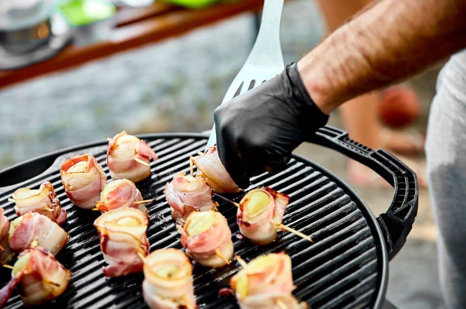 Close-up of a person's hand wearing a black glove, using a grilling spatula to cook skewers wrapped in bacon on a round charcoal grill.