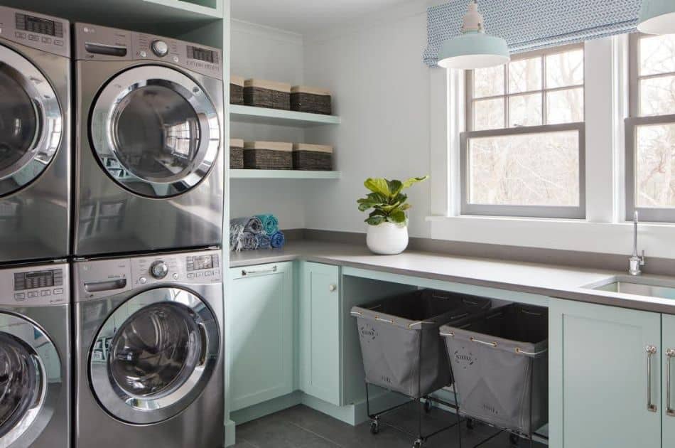 A modern and tidy laundry room with light teal cabinetry and gray countertops. There are two sets of stainless steel washer and dryer units stacked on the left. On the right, there is a deep sink beneath a window with a woven blue window shade, and a potted green plant adds a touch of freshness to the space. Above the sink, open shelves hold baskets and folded towels, while two labeled laundry hampers on wheels sit neatly underneath the counter.