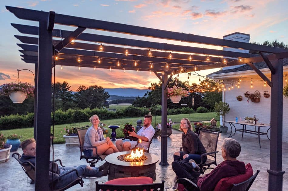 A group of people relaxing on a patio at dusk, with a fire pit in the center providing a cozy atmosphere. String lights are strung overhead, casting a warm glow on the scene. In the background, a breathtaking sunset paints the sky in shades of pink and orange, with mountains silhouetted on the horizon. The patio is adorned with potted plants and hanging baskets, creating an inviting outdoor living space.