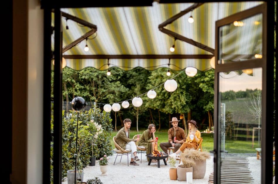 View from inside a house looking out to a stylish outdoor gathering space under a striped awning. A group of four people is seated around a small fire pit, engaged in conversation, with string lights above adding a warm glow to the evening.