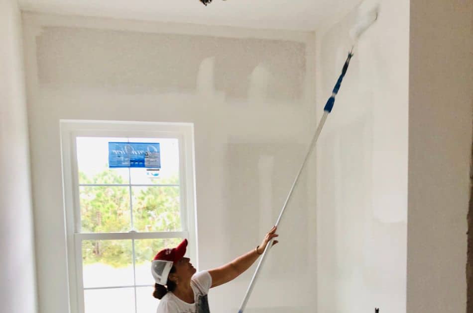 A woman is painting a wall with a long-handled roller brush, reaching up towards the ceiling.