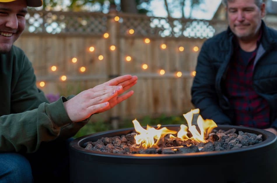 An outdoor setting featuring two people enjoying a fire pit during twilight. The focus is on a smiling man in the foreground extending his hands towards the warmth of the flames, with an out-of-focus companion in the background. String lights are visible on a fence behind them, adding a cozy ambiance.
