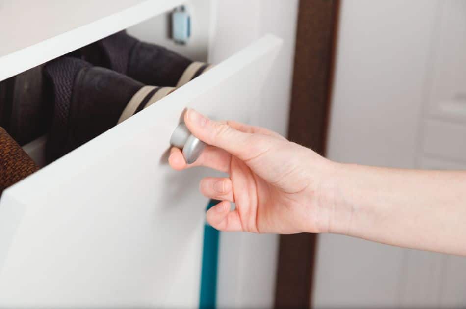 A person's hand is pulling open a white laundry drawer with a metallic handle, revealing dark-colored shoes inside.