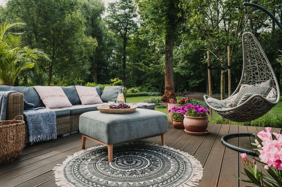 An inviting garden patio with a large, comfortable corner sofa adorned with cushions in shades of gray and soft pink. A stylish hanging egg chair provides a unique seating option. The area features a decorative round rug and a low, central ottoman with a tray of grapes on top.