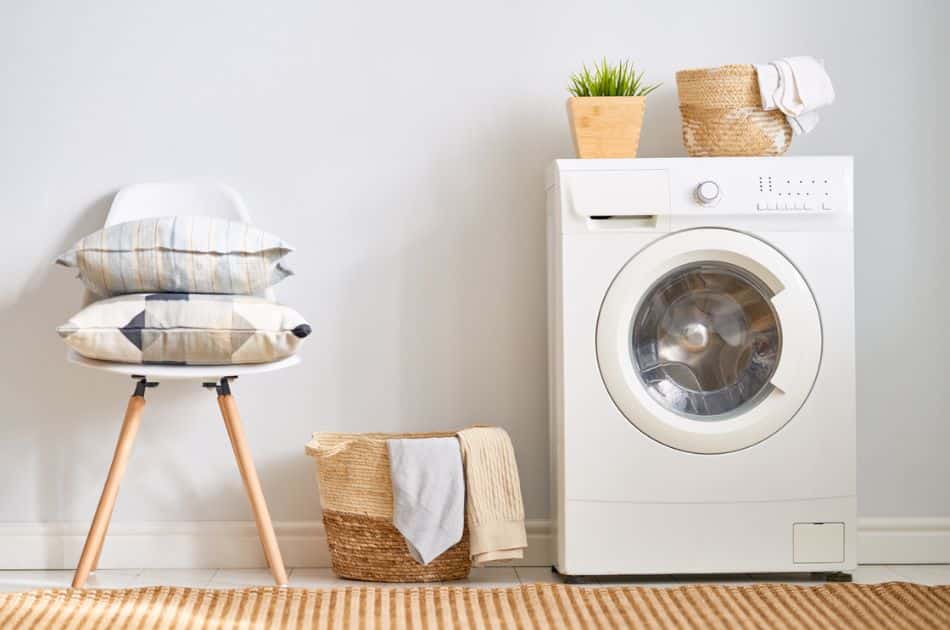 A simple and clean laundry room corner with a modern white front-loading washing machine on the right. On top of the washer, there is a small potted plant and a woven basket with towels. To the left is a white chair with wooden legs, stacked with two patterned cushions, and beside it, a woven basket holds a folded blanket. The room has a neutral color palette, complemented by a natural fiber rug on the floor.