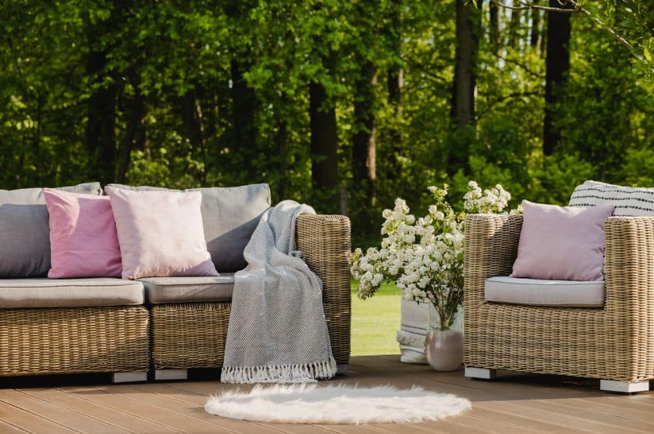 A cozy outdoor patio area with wicker furniture adorned with soft grey and pastel pink cushions. A grey throw blanket casually draped over one of the chairs adds a homey touch. In the center, a vase with white blossoms brings a fresh and inviting feel to the space. The setting is on a wooden deck with a lush green forest in the background.