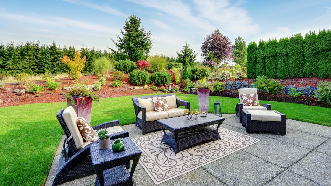 An elegant outdoor living space with modern black rattan furniture, including a sofa, armchairs, and a coffee table, set on a patterned area rug. The area is decorated with cushions and a vase with flowers, enhancing the comfort and aesthetic appeal. The patio overlooks a beautifully landscaped garden with a variety of lush trees and shrubs, bordered by a tall privacy hedge under a clear blue sky.