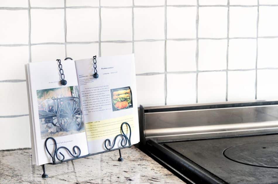 A cookbook is open on a decorative metal stand with intricate scrollwork, set against a kitchen counter with a subway tile backsplash. The page displays a colorful recipe with images, held in place by elegant black chains with clips.