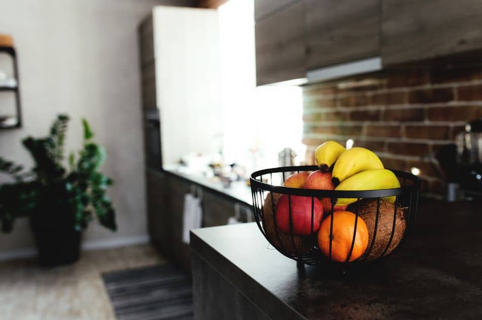 A contemporary kitchen with a rustic touch features a black wire fruit bowl on a dark countertop. The bowl is filled with an assortment of ripe fruit, including bananas, apples, an orange, and a coconut, offering a pop of color against the muted tones of the room.