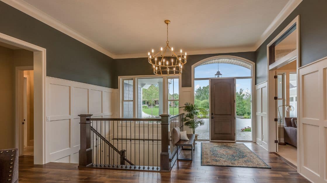 An elegant, well-lit entryway with a dark wood floor, contrasting white wainscoting, and dark gray walls. An open front door with arched transom windows allows natural light to flow in, highlighting the chandelier above.