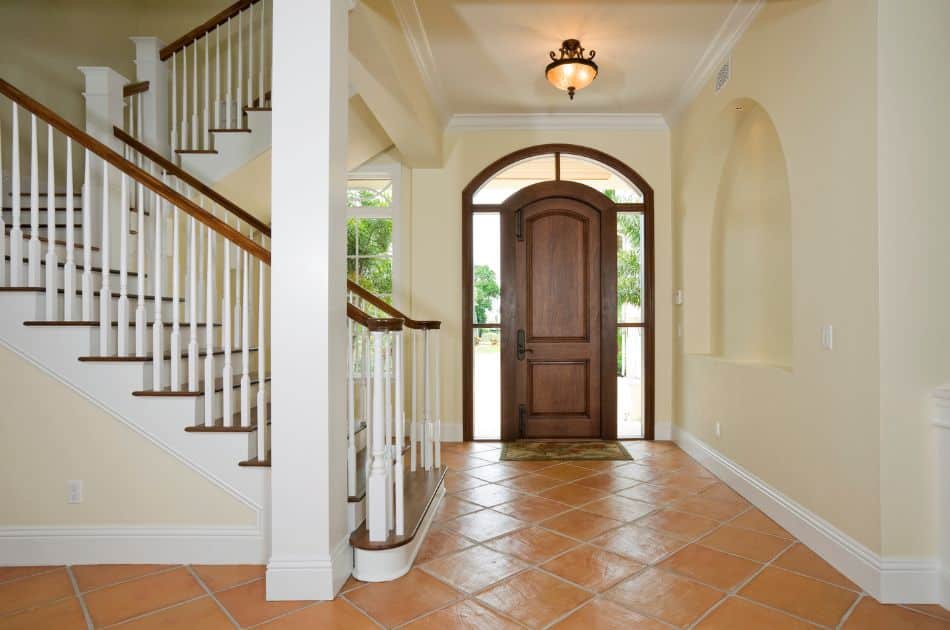 A spacious and inviting foyer featuring terra cotta tiled flooring and a grand staircase with white spindles and polished wooden handrails. An arched wooden front door with glass panels and a semi-circular transom window opens to reveal a glimpse of a lush outdoor view. The walls are painted in a soft beige tone, with an alcove niche and white molding adding architectural interest. A classic ceiling light fixture provides a warm ambiance to the space.
