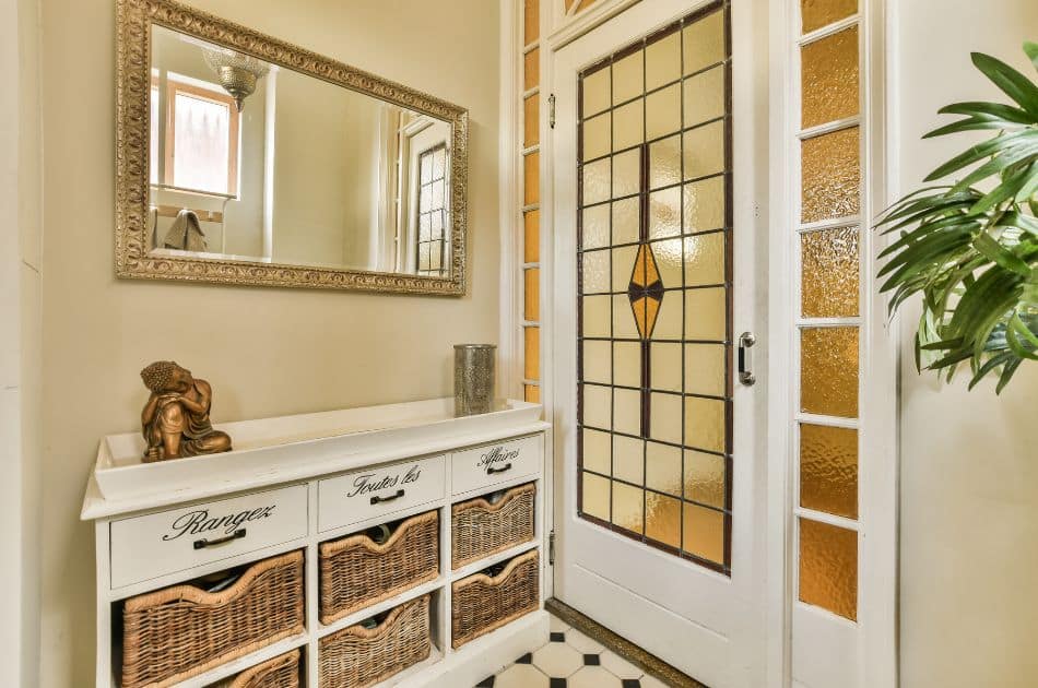 A quaint entryway featuring a white vintage-style console table with labeled drawers and wicker baskets. Above it hangs an ornate mirror reflecting a wall-mounted light fixture. To the right, a door with a stained glass window incorporating yellow tones and a central diamond-shaped design adds a touch of color and privacy. Beside the door, a lush green plant adds a touch of life to the space. The floor is tiled with a classic black and white pattern.