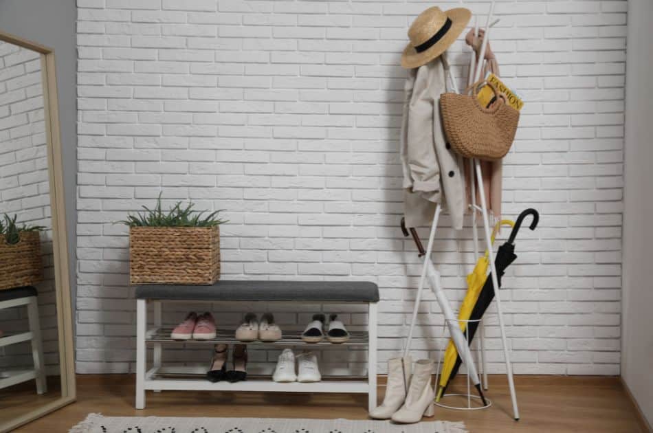 A modern and minimalist entryway with a white brick wall. On the left, a full-length mirror reflects a woven basket planter with greenery, positioned atop a wooden shelf. In the center, a bench with a gray cushion provides seating, with an array of shoes neatly organized below. To the right, a stylish white coat rack holds a straw hat, a beige trench coat, a woven tote bag with a magazine, and a yellow umbrella, adding a pop of color. The scene is completed by a patterned rug on the wooden floor.