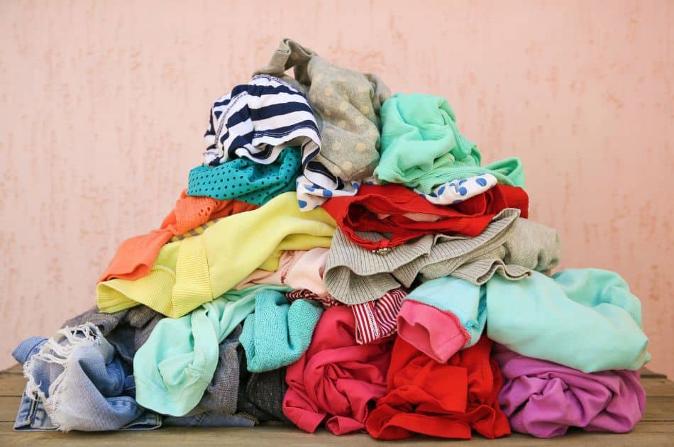 A large, colorful pile of assorted clothes sits on a surface against a peach-colored wall with a subtle texture. The clothing items include jeans, shirts, and sweaters in a variety of patterns and colors.