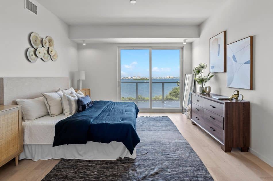 A serene bedroom with a waterfront view through a large floor-to-ceiling window. The bed is dressed in white linens with a textured pattern and topped with a dark blue throw, accompanied by neutral-toned pillows. The light-colored upholstered headboard matches the bed’s wooden frame. To the left, an artistic arrangement of decorative plates adorns the wall. On the right, a dark wood dresser is accessorized with a small plant, vases, and two framed abstract art pieces, adding a modern touch to the space. A shaggy dark grey area rug lies on the floor, complementing the lighter hardwood flooring.