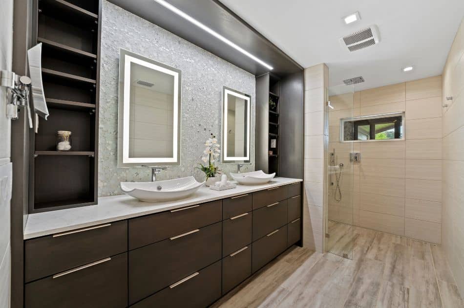 A luxurious and modern bathroom with a spacious layout. The vanity features dark wood cabinets with sleek, horizontal handles and a white countertop. Two vessel sinks are positioned beneath illuminated rectangular mirrors mounted on a textured tile backsplash. To the left of the vanity is an open shelving unit for storage. The large walk-in shower on the right has light beige tiles, a built-in niche for toiletries, and a glass partition. The shower area includes a rainfall showerhead and a handheld showerhead. The flooring is a light wood-look tile, adding warmth to the contemporary design. Recessed lighting and a ventilation fan are installed in the ceiling. An orchid plant and decorative items on the vanity add a touch of elegance.