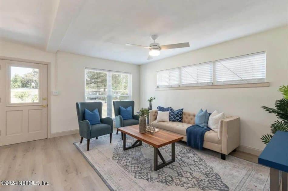 A bright living room with off-white walls and light wood flooring. The room features a beige tufted sofa with blue and beige throw pillows, accompanied by a blue throw blanket. In front of the sofa is a wooden coffee table with metal legs, decorated with plants and decorative items. Two teal armchairs with blue cushions are positioned near a large window on the left, which lets in natural light. Above the sofa are three narrow horizontal windows with blinds. A ceiling fan with a light fixture is centrally located on the ceiling. The entrance door, with a glass insert, is to the left of the window. A patterned area rug anchors the seating area. A green potted plant is visible in the right corner.