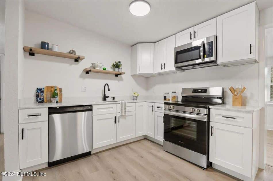 A modern kitchen with white cabinets and light wood flooring. The cabinetry features black handles and includes a mix of drawers and cupboards for ample storage. Stainless steel appliances include a dishwasher, a stove with an oven, and an over-the-range microwave. The countertops are white, providing a clean and bright workspace. Above the countertops, two wooden floating shelves are mounted on the wall, displaying decorative items like mugs, bowls, and small plants. The sink area includes a black faucet and a striped dish towel. A ceiling light fixture provides illumination, and a window near the stove allows natural light to enter.