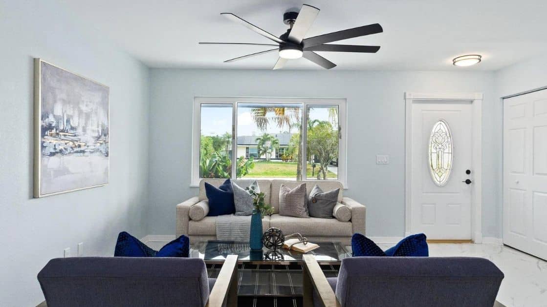 A modern living room with light blue walls, a large window, and a stylish ceiling fan. The room features a comfortable beige sofa adorned with a mix of grey and navy throw pillows, positioned against the window. In front of the sofa is a glass coffee table with decorative items. Two navy chairs are partially visible in the foreground. A piece of abstract artwork hangs on the left wall. The entrance door, with an oval glass insert, is situated to the right of the window. The room is bright and airy, with a view of a lush green yard outside the window.