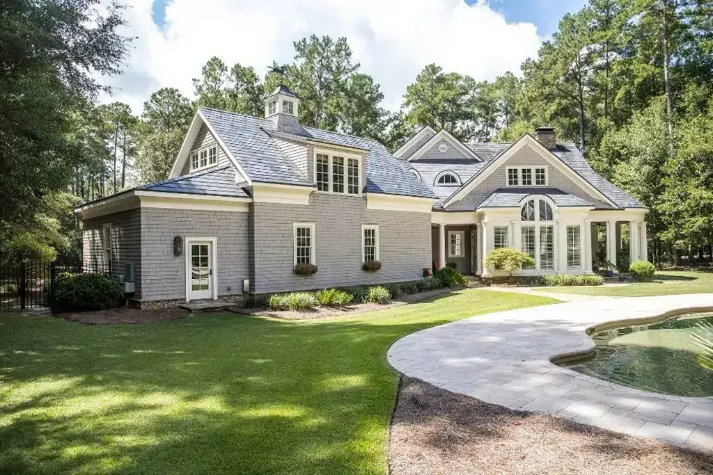 A charming Cape Cod-style house featuring grey shingle siding, white trim, and multiple gabled dormer windows. The house has a large, steeply pitched roof with a central cupola. There are well-maintained shrubs and plants around the house, and a paved patio area leading to a pool with an irregular shape. The backdrop consists of tall, mature trees, adding to the serene and picturesque setting. The house has large, elegant windows and a covered entryway supported by white columns. house style names
