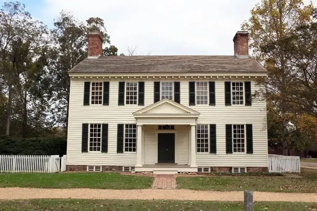 A classic Colonial-style house with a symmetrical facade and light yellow clapboard siding. The house features two brick chimneys on either side of the roof, black shutters on the windows, and a central entryway with a pediment supported by columns. The front door is painted black and flanked by sidelights, with a small brick walkway leading up to it. The house is surrounded by a white picket fence, and the front yard is a simple, grassy lawn with trees in the background, adding to the historic and charming appearance.
