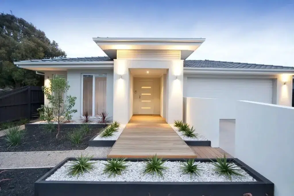 A sleek and modern contemporary-style house with a white exterior and clean lines. The entrance features a minimalist design with a flat roof and a recessed doorway, illuminated by recessed lighting. The front door is stylish with horizontal glass panels. A wooden walkway leads to the entrance, flanked by white stone landscaping with low-maintenance plants and shrubs. The house includes a large window and an attached garage with a white door. The front yard is designed with a combination of dark mulch, white stones, and neatly arranged plants, creating a sophisticated and low-maintenance landscape. The overall atmosphere is clean, modern, and inviting.