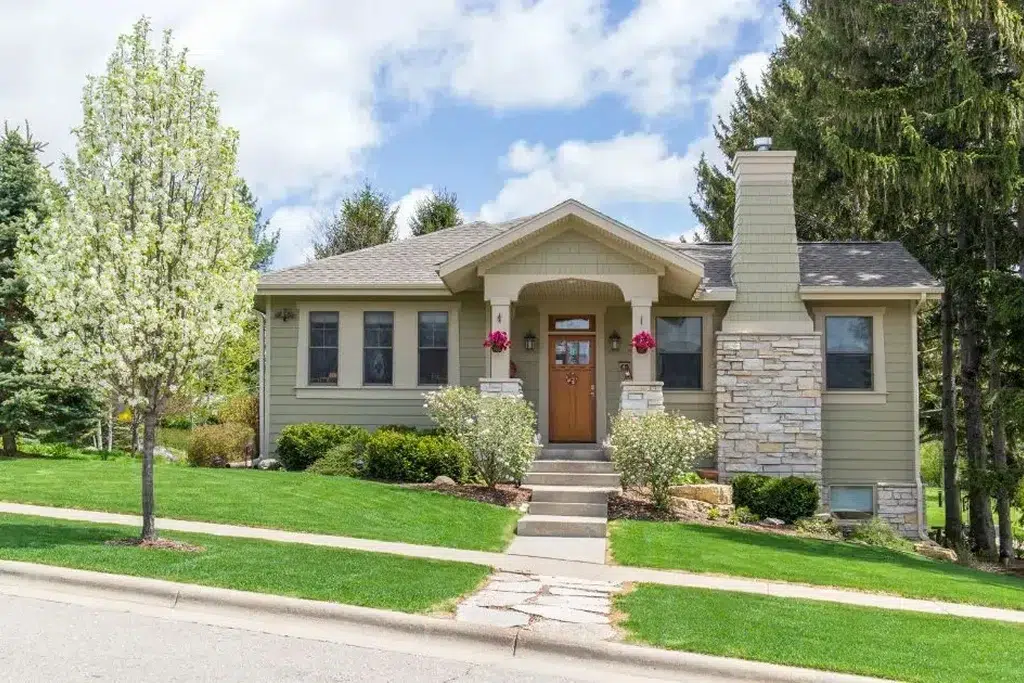 A charming Craftsman-style house with a light green exterior and stone accents. The house features a covered front porch with tapered columns and decorative brackets, and a wooden front door with glass panels. The windows have white trim and are symmetrically placed on either side of the porch. The front yard is beautifully landscaped with green grass, flowering shrubs, and a blossoming tree. A set of concrete steps leads up to the porch from the sidewalk.