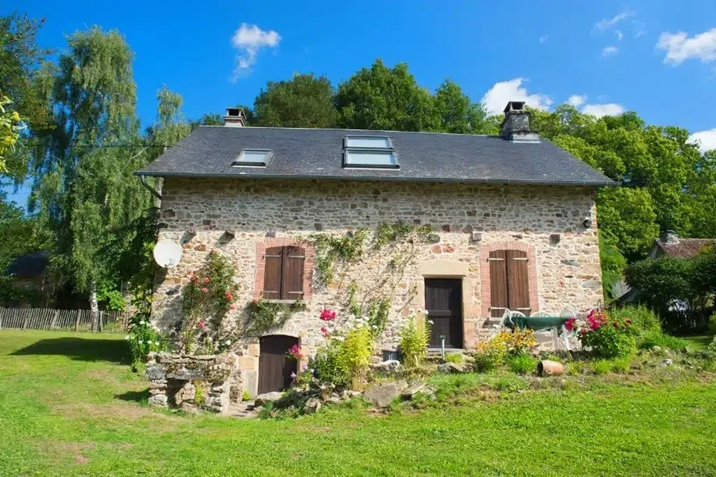 A quaint French country home with rustic stone walls and a dark slate roof. The house features wooden shutters on the windows and a charming front door surrounded by climbing vines and flowering plants. The upper floor has a skylight window, and a satellite dish is attached to the side of the house. The front yard is lush and green, with a variety of colorful flowers and shrubs adding to the idyllic countryside setting. Tall trees and a bright blue sky with a few scattered clouds provide a picturesque backdrop, enhancing the home's peaceful and pastoral ambiance.