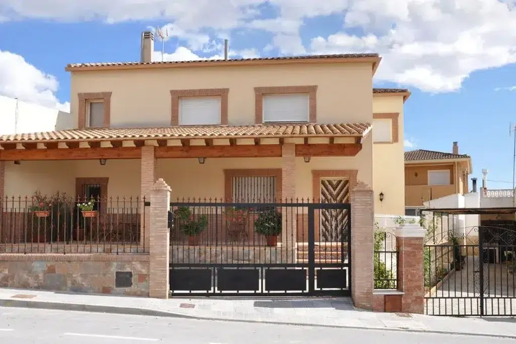 A Mediterranean-style home with warm beige stucco walls and a terracotta tiled roof. The house features large windows with decorative brick trim and a covered porch with wooden beams and brick columns. The front yard is enclosed by a wrought-iron fence with brick pillars, and potted plants add greenery and charm to the entrance. There is a gated driveway on the right side leading to a garage, and the sky above is dotted with fluffy white clouds, giving the scene a bright and sunny atmosphere.