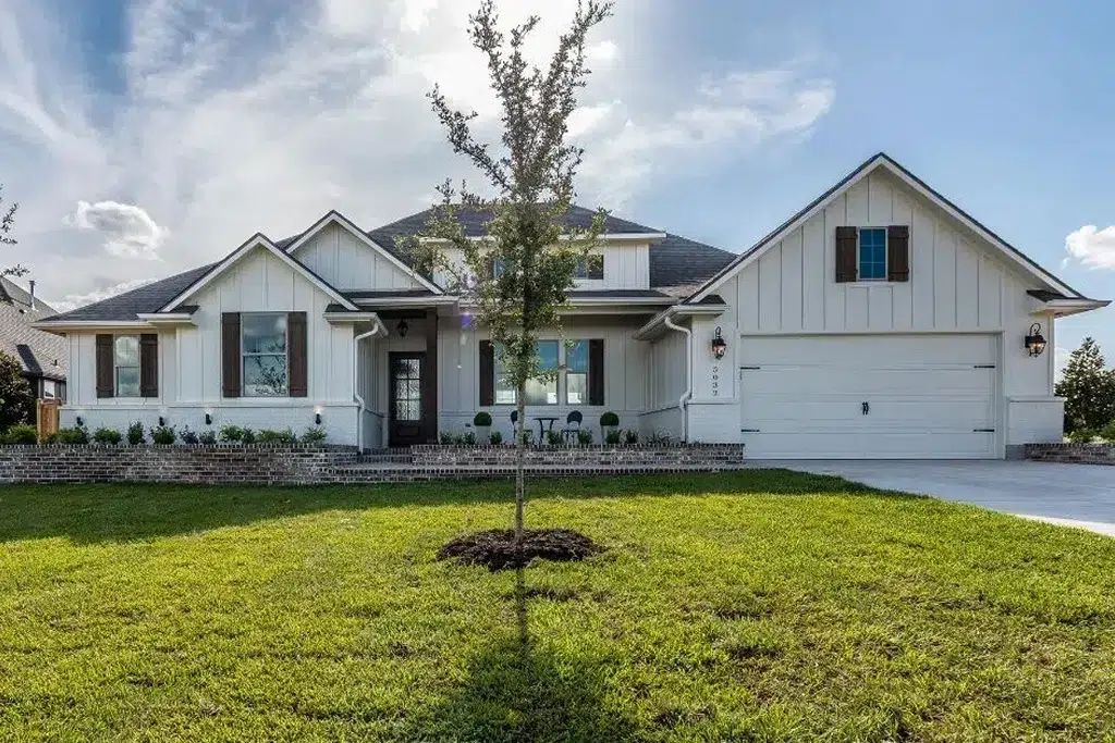 A modern farmhouse-style house with a white exterior and dark trim, featuring a combination of board-and-batten siding and brick accents. The house has a spacious front porch with black wrought iron railing and wooden shutters on the windows. The front yard is landscaped with a well-manicured lawn, small shrubs, and a young tree planted in the center. The house includes a large, attached garage with carriage-style doors. The sky is partly cloudy, and the overall atmosphere is bright and inviting.
