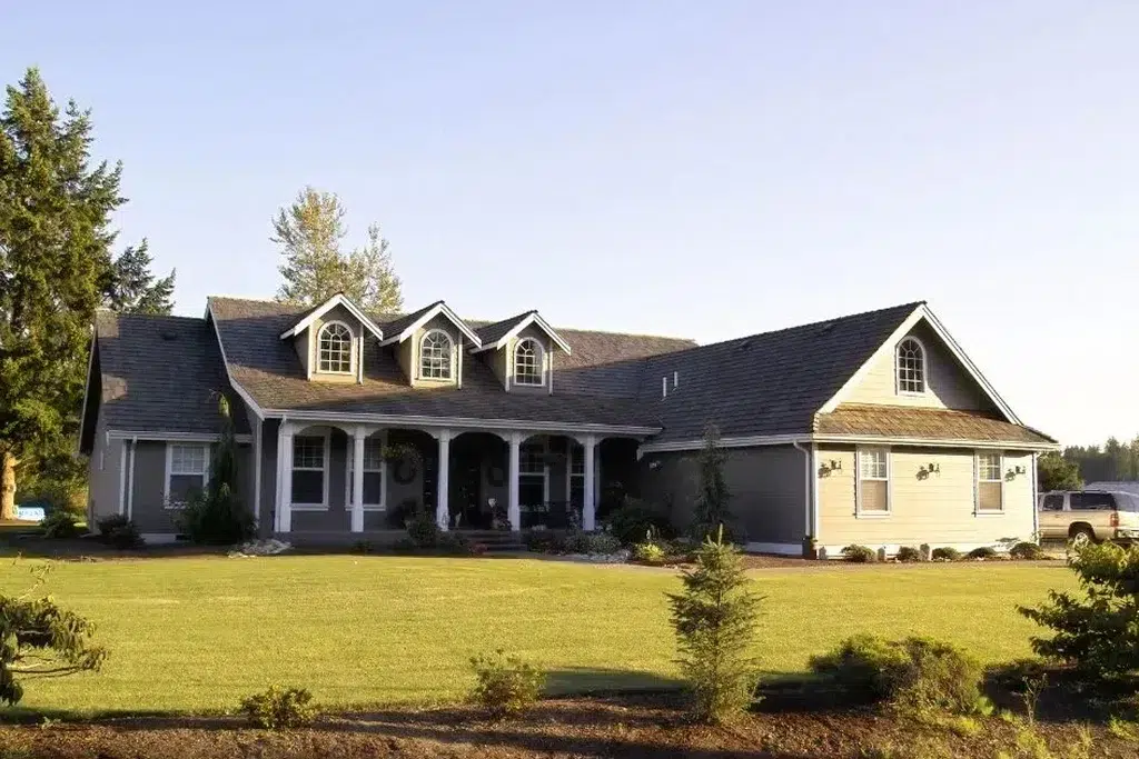 A modern ranch-style house with grey siding and white trim, featuring a large, sprawling single-story layout with multiple gabled dormer windows. The house has a welcoming front porch with white columns and a well-maintained garden with shrubs and flower beds. The spacious front yard is landscaped with a lush green lawn and a few young trees, and a pickup truck is parked on the driveway to the right. The backdrop includes tall trees and a clear sky, giving the scene a serene and picturesque feel.
