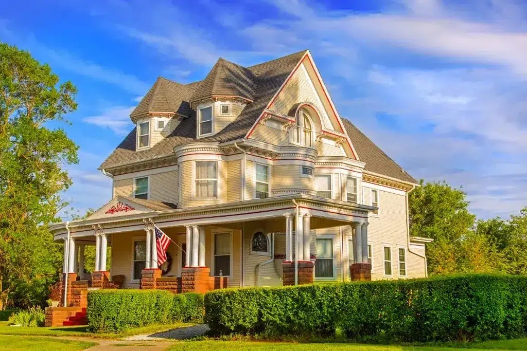 A grand Victorian-style house with a light grey exterior and intricate architectural details. The house features a steeply pitched roof with multiple gables, a prominent rounded turret, and a large front porch supported by white columns and red brick bases. Decorative trim and accents are painted in white and red, adding to the elaborate design. The front yard is neatly landscaped with hedges and a lush green lawn. An American flag is displayed near the entrance, and the sky above is a vibrant blue with some scattered clouds, creating a picturesque and timeless scene.