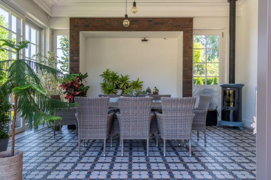 A bright dining area with large windows allowing plenty of natural light. The room features a patterned tile floor, a brick accent wall, and a cozy wood-burning stove. A wicker dining set with eight chairs surrounds a large table, and various potted plants add a touch of greenery to the space.