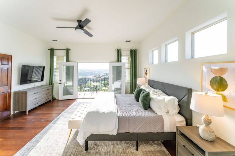 home staging lighting: A spacious and bright bedroom featuring a large bed with gray bedding and green accent pillows. The room has a modern aesthetic with clean lines and minimalistic decor. Large windows above the bed and French doors leading to a balcony let in plenty of natural light. The room includes a ceiling fan, a flat-screen TV mounted on the wall, a dresser, and two nightstands with lamps. The floor is covered with a light-colored rug over hardwood flooring, and the walls are painted white, enhancing the airy feel of the space.