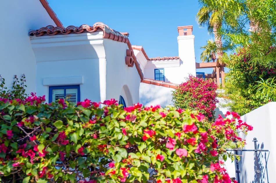 A Mediterranean-style house with white stucco walls and terracotta roof tiles, featuring blue-framed windows and doors. The exterior is adorned with vibrant bougainvillea plants in full bloom, adding splashes of pink and red.