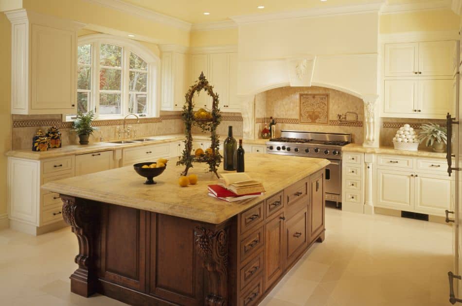 modern mediterranean home design ideas: A Mediterranean-style kitchen with cream-colored cabinets and a large central island featuring intricate woodwork. The countertops are made of light-colored stone, and the backsplash has decorative tile work. The kitchen includes a professional-grade stainless steel stove with an ornate range hood. A large arched window above the sink allows natural light to fill the room. Decorative elements, such as a fruit arrangement and wine bottles, are placed on the island, adding to the elegant and welcoming atmosphere.