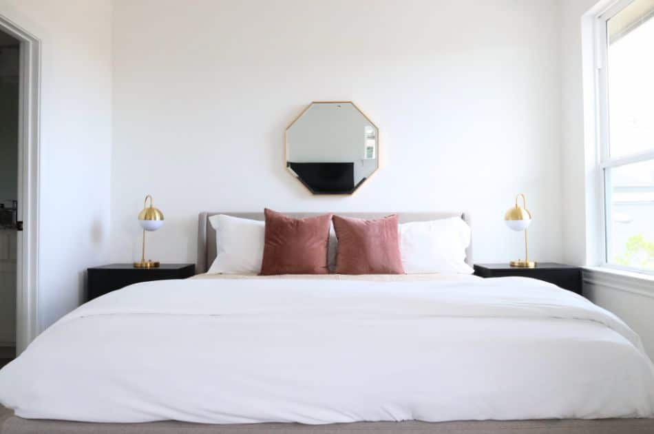 A contemporary bedroom with a minimalist design featuring a large bed with white bedding and two pink accent pillows. The bed is flanked by black nightstands, each adorned with a modern gold and white table lamp. Above the bed hangs a geometric octagonal mirror, adding a stylish touch to the room. Natural light streams in through a window on the right, illuminating the clean white walls and creating a bright, inviting atmosphere. The overall decor is simple yet elegant, emphasizing comfort and modern aesthetics.