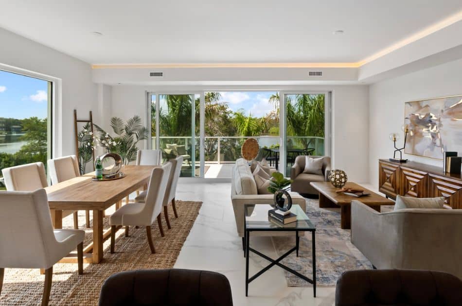 A spacious, contemporary living and dining area featuring large windows that let in abundant natural light. The room has a minimalist design with white walls and white marble flooring. The dining area includes a wooden table with eight beige upholstered chairs on a woven jute rug.