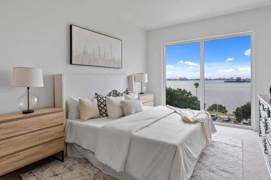 A serene bedroom with a minimalist design, featuring a large window offering stunning views of a lake and city skyline. The room is decorated in neutral tones with a white upholstered bed adorned with a mix of white and black geometric-patterned pillows. Two wooden nightstands with matching glass-base lamps flank the bed. A light beige area rug covers the floor, and the room is accented with abstract wall art above the bed.