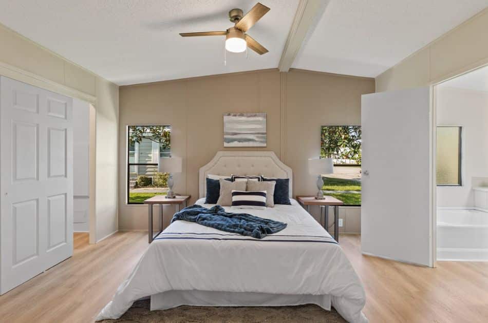 A cozy bedroom with a neutral color palette, featuring a queen-sized bed with a tufted headboard and crisp white bedding accented by navy blue and striped pillows. At the foot of the bed is a neatly folded blue throw blanket. The room is well-lit by natural light coming through two windows on either side of the bed for condo staging.