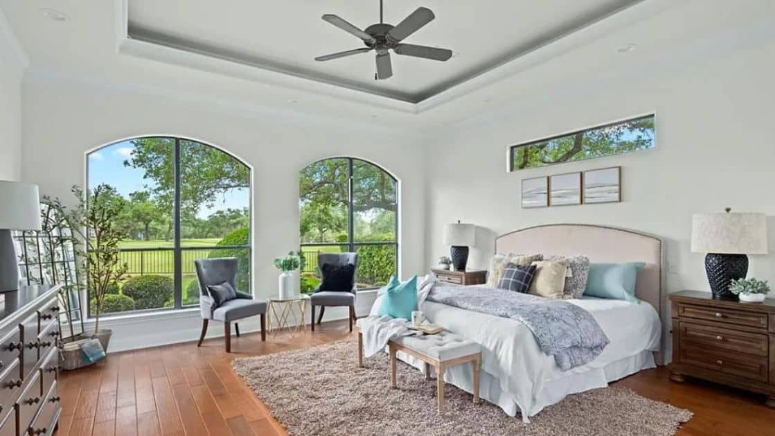 This image features a contemporary master bedroom with a calm and inviting ambiance. The room is spacious, with large, arched windows that allow ample natural light to flood the space, offering a serene view of the greenery outside. The centerpiece of the room is a cozy bed with a tufted headboard, dressed in soft, neutral-toned bedding, complemented by a variety of pillows in different textures and patterns.