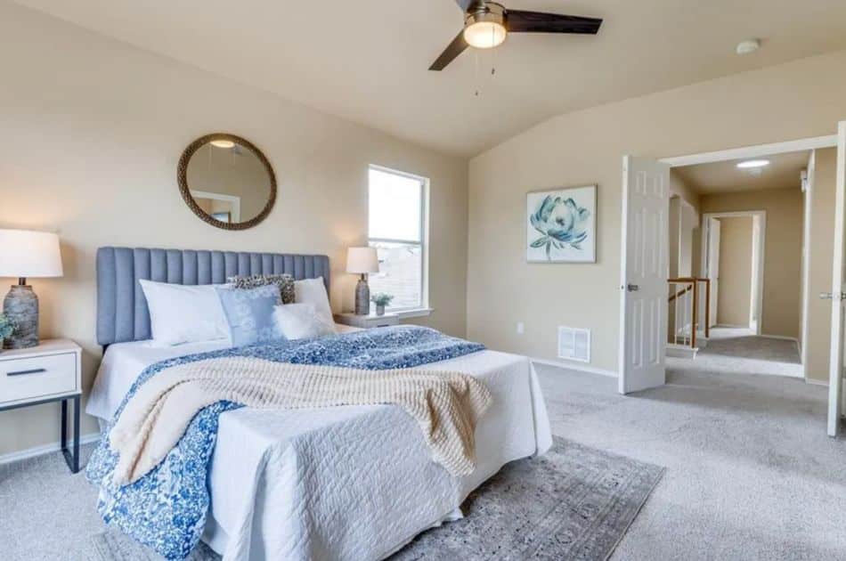 A cozy bedroom with soft, neutral tones featuring a large bed with a tufted gray headboard. The bed is dressed in white bedding with blue accents and a cream-colored throw blanket. On either side, there are matching nightstands with decorative lamps. Above the bed, a round mirror adds a stylish focal point. A ceiling fan hangs above, and natural light streams in through a window. The room is carpeted and opens up to a hallway, with a floral artwork on one wall adding a touch of color.