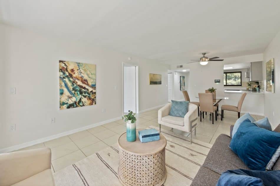 A bright and inviting living room with an open layout extending into the dining area. The space features a comfortable seating arrangement, including a modern gray sofa adorned with blue and gray throw pillows, and a cozy beige armchair with a blue cushion.