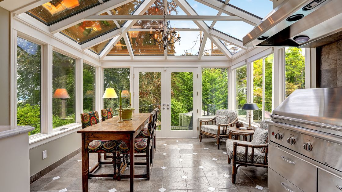 The image shows a beautifully designed Florida Room with large glass windows and a slanted glass ceiling, allowing natural light to flood the space. The room features a cozy dining area with an ornate wooden table and colorful cushioned chairs. Adjacent to the dining table, there is a comfortable seating area with patterned armchairs and small side tables. A built-in stainless steel grill adds functionality to the space, perfect for indoor-outdoor cooking.