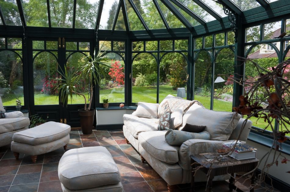 The image shows an elegant Florida Room with dark green framing and floor-to-ceiling windows that overlook a lush, well-maintained garden. The space is furnished with plush, cream-colored sofas and matching ottomans, creating a comfortable and cozy seating area.