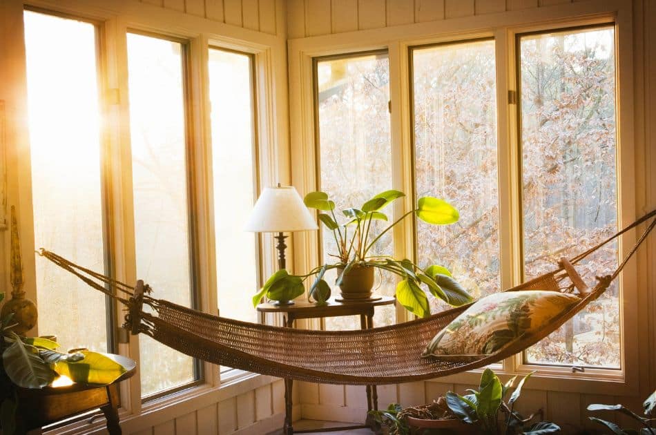 The image shows a tranquil Florida Room with large windows that allow warm sunlight to pour in, creating a peaceful, sun-drenched environment. A woven hammock stretches across the room, complete with a decorative pillow, making it a perfect spot for relaxation. A small table next to the hammock holds a potted plant and a table lamp, adding a touch of greenery and a cozy ambiance to the space | how to decorate a florida room