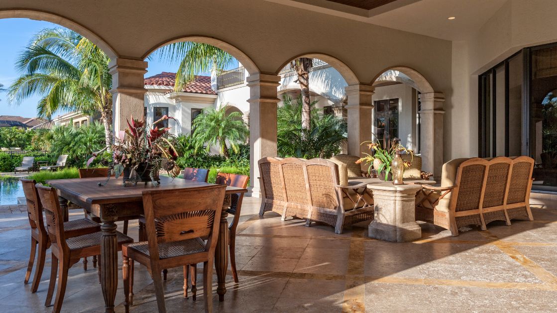 The image shows a luxurious outdoor lanai featuring a dining table with wooden chairs and a sitting area with woven chairs around a stone table. The space is covered by a series of elegant arched columns, creating a Mediterranean-style vibe. The lanai overlooks a lush garden with tropical plants, palm trees, and a pool, set against the backdrop of a large home with a terracotta-tiled roof.