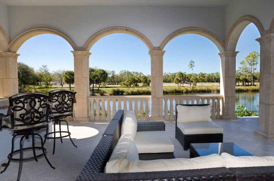 The image shows a serene and elegant lanai with arched columns framing a view of a tranquil waterfront and lush greenery. The seating area features a modern wicker sectional with white cushions and a glass-topped coffee table, creating a cozy spot to relax.