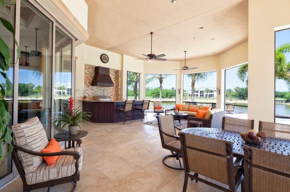 The image shows a stylish and spacious lanai with an outdoor kitchen and a seating area. The kitchen features a stone backsplash, a grill with a chimney-style hood, and a bar counter with stools. The lanai is furnished with comfortable cushioned chairs and a dining table, complemented by ceiling fans for added comfort.