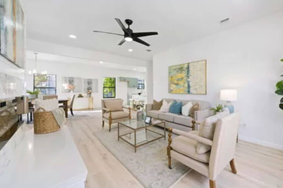 The image shows a bright, open living room and dining area with a modern yet cozy aesthetic. The living room features a neutral-toned sofa with blue and beige accent pillows, two beige armchairs, and a glass coffee table on a light area rug.